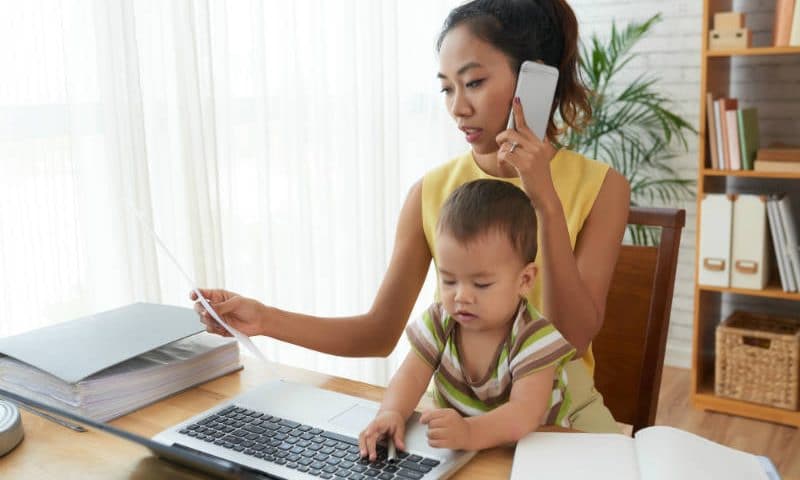 a mom works in front of a computer, on the phone with a document in her hand and a baby on her lap